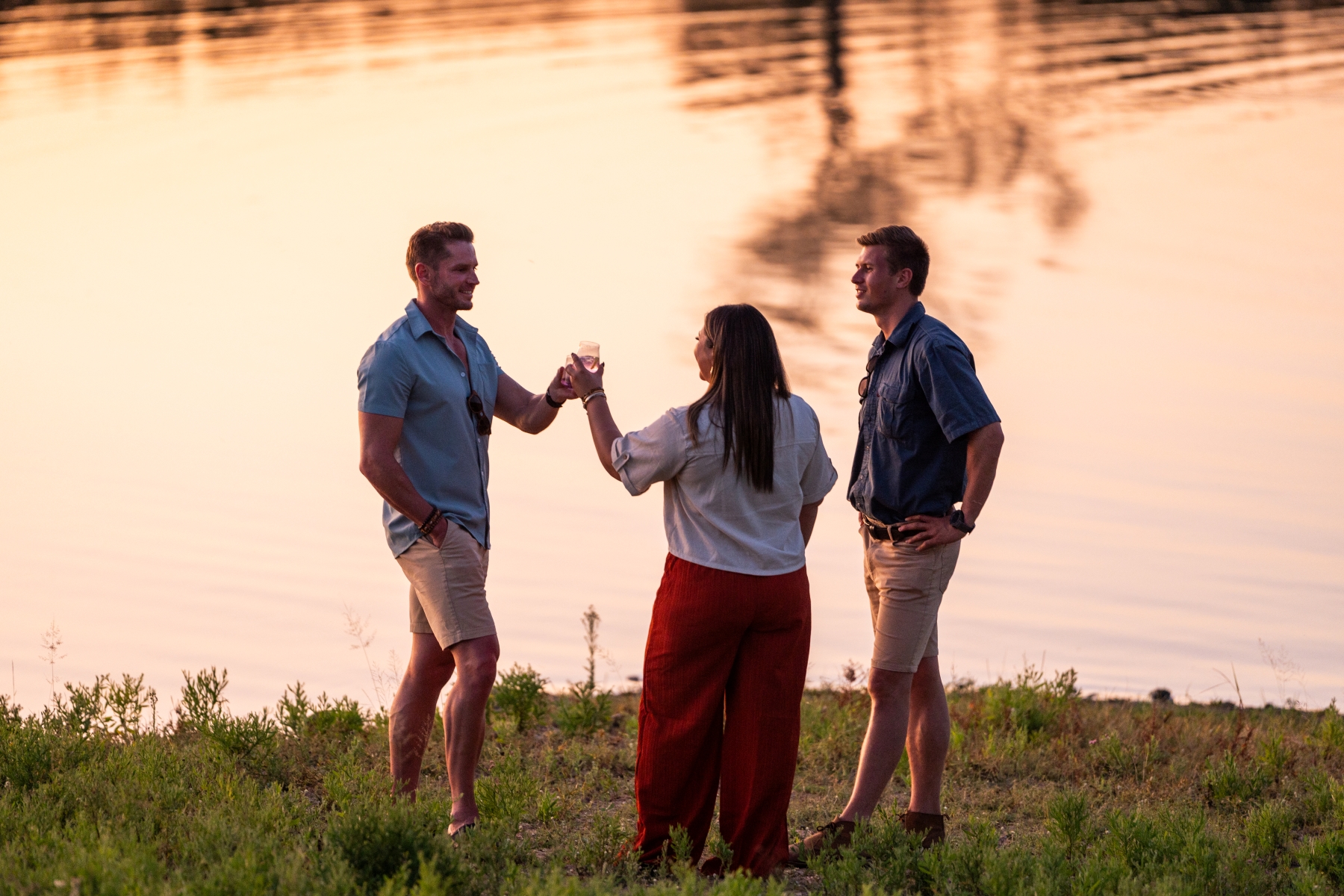 two-guests-stand-with-their-guide-on-the-waters-edge-raising-sundowners-as-the-setting-sun.jpg