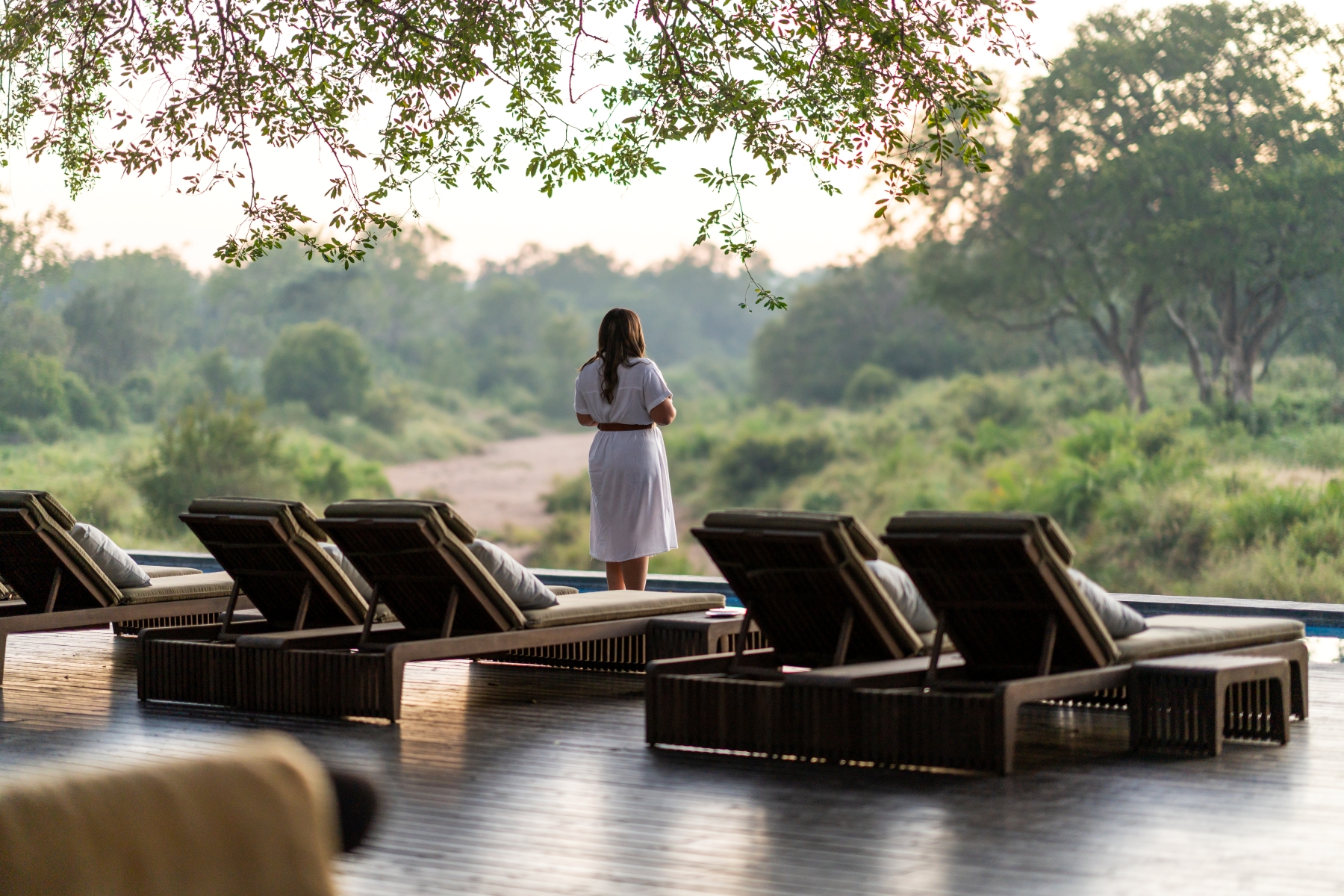 a-woman-in-a-white-dress-stands-facing-the-bush-beyond-pool-loungers.jpg