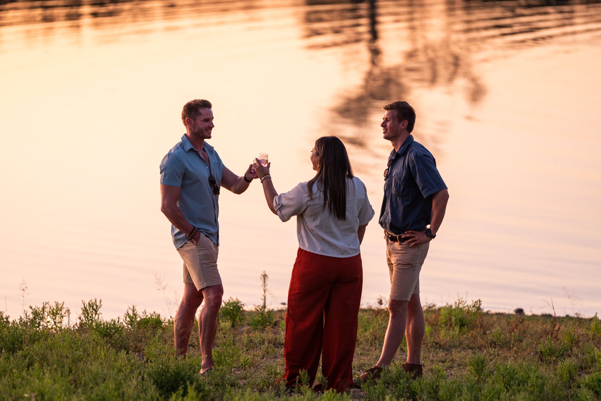 Three safari-goers enjoy a sunset drink by the water’s edge, toasting to an unforgettable African adventure.